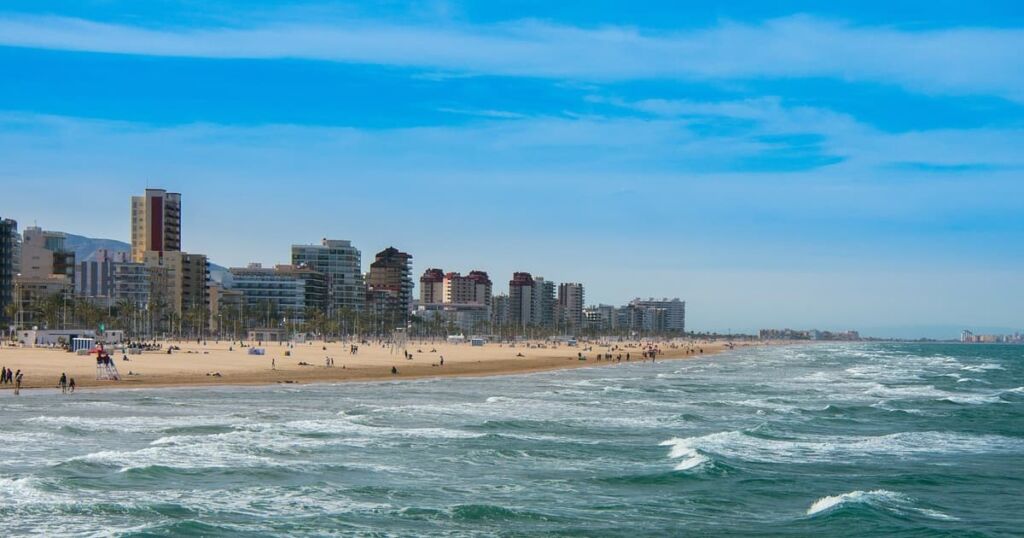 Agence immobilière à Gandia Vue de la mer agitée à Gandia, avec des vagues qui s'échouent sur la plage, la ville visible en arrière-plan, et un ciel bleu turquoise vibrant.