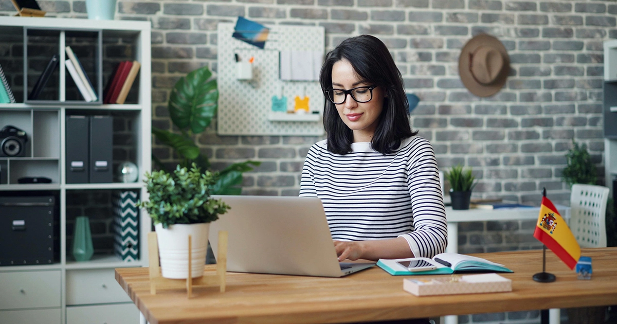 femme_préparant_son_irpf sur son ordinateur Femme assise devant son ordinateur portable avec un drapeau espagnol posé sur le bureau