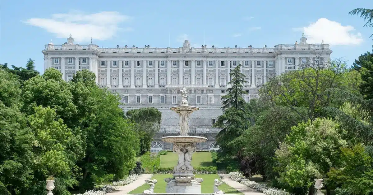 Le Palais Royal de Madrid, vu depuis ses jardins formels, avec une fontaine centrale ornée de statues et une végétation luxuriante autour.