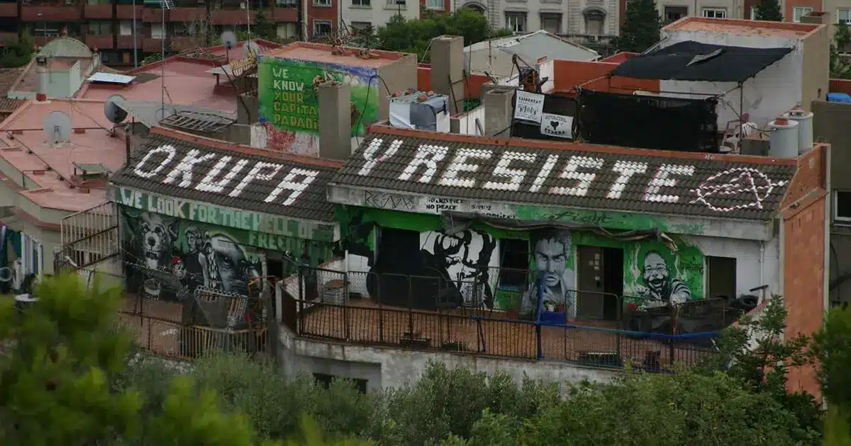 Okupas Toit d’un bâtiment occupé à Barcelone avec des graffitis militants, notamment le slogan « Okupa y resiste » (« Occupe et résiste »), illustrant le mouvement des squatters en Espagne.
